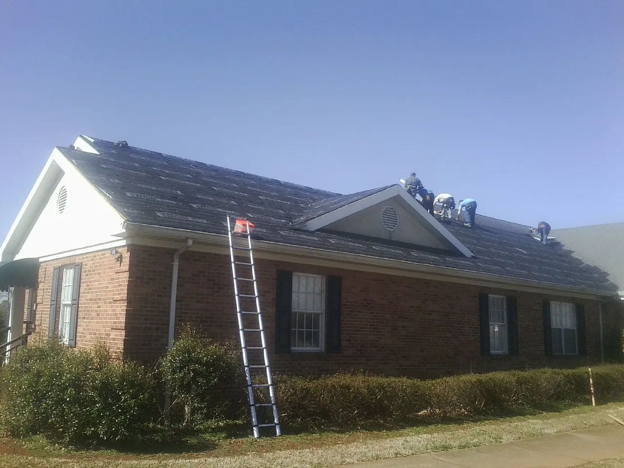 Crew tearing shingles off a brick ranch roof with a ladder set — Metal Roof Repair work by our crew in Chardon