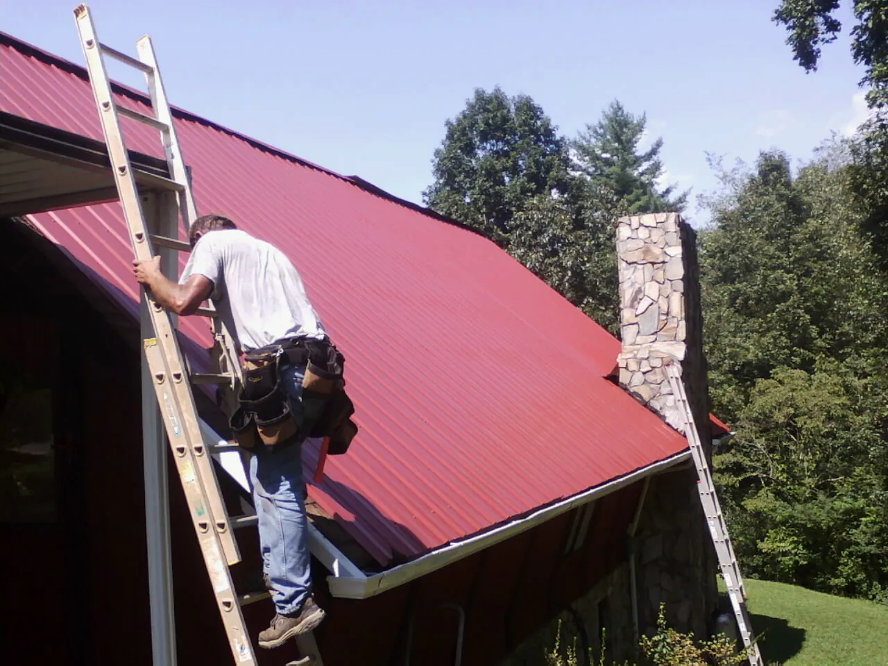 Roofer on a ladder beside a red metal roof and stone chimney — Soffit & Fascia Repair by Roofing Brothers in Chardon